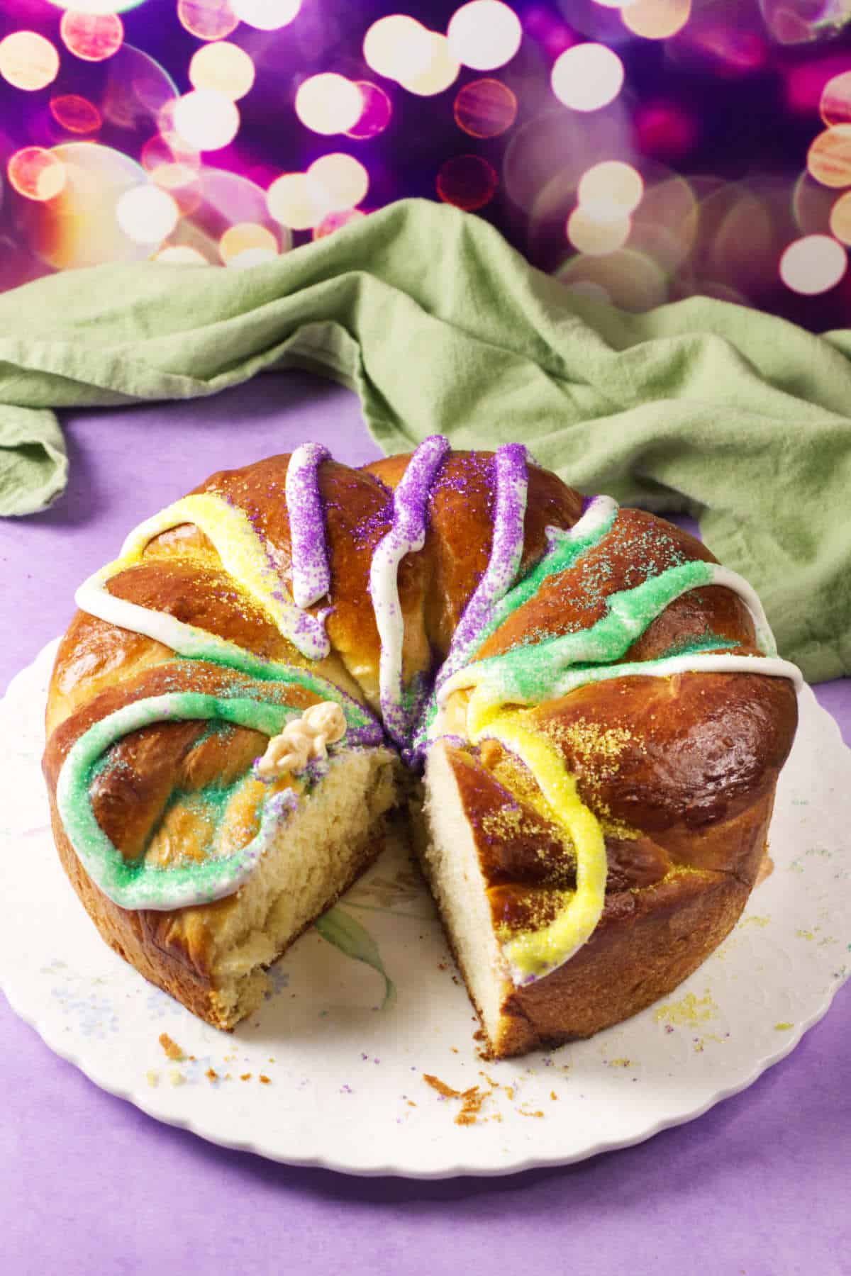 A round Mardi Gras king cake decorated with colorful purple, yellow, and green sugar and icing sits on a white plate with a slice cut out. A green cloth and festive blurred lights are in the background.