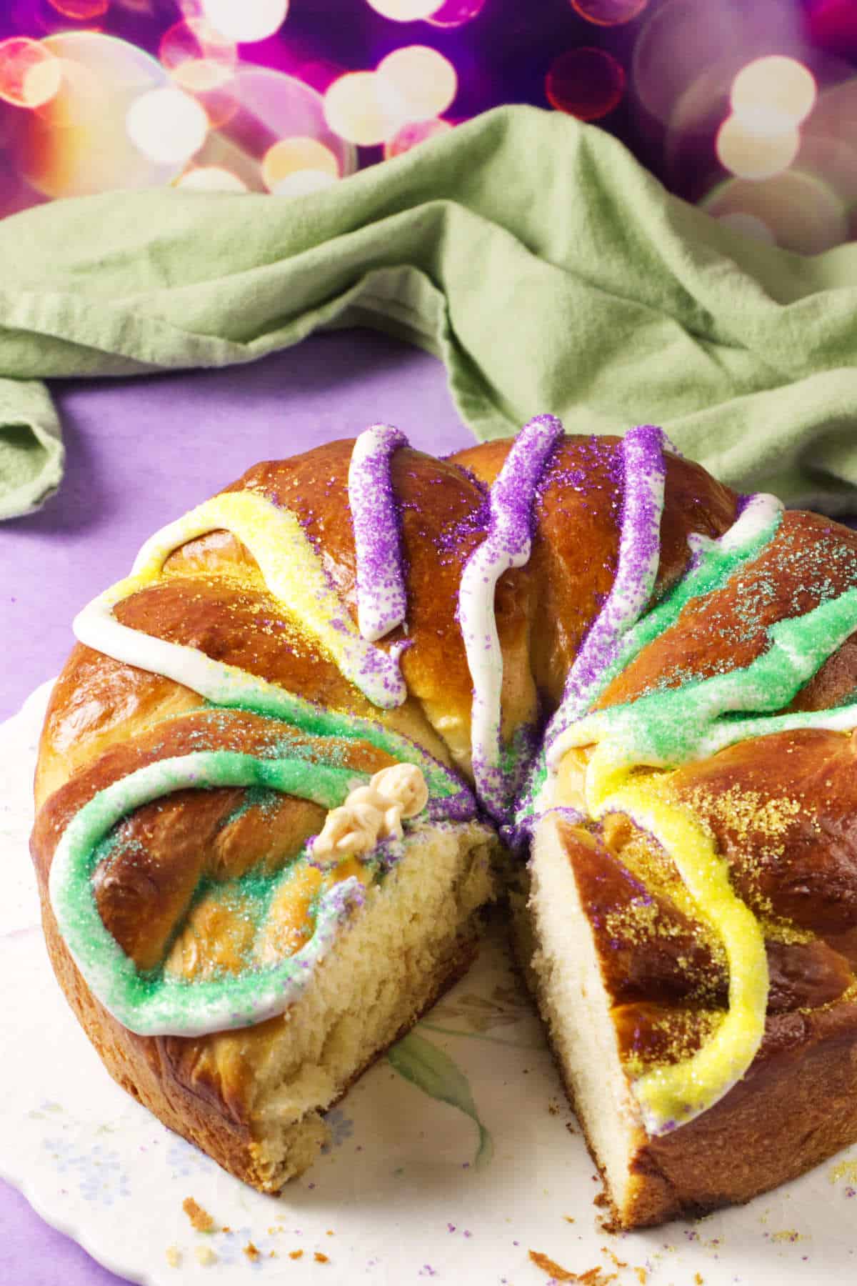 A round King Cake decorated with green, purple, and yellow icing and sugar sits on a white plate. One slice has been cut, revealing the soft, fluffy interior—perfect for celebrating Mardi Gras. A green cloth and colorful lights are in the background.