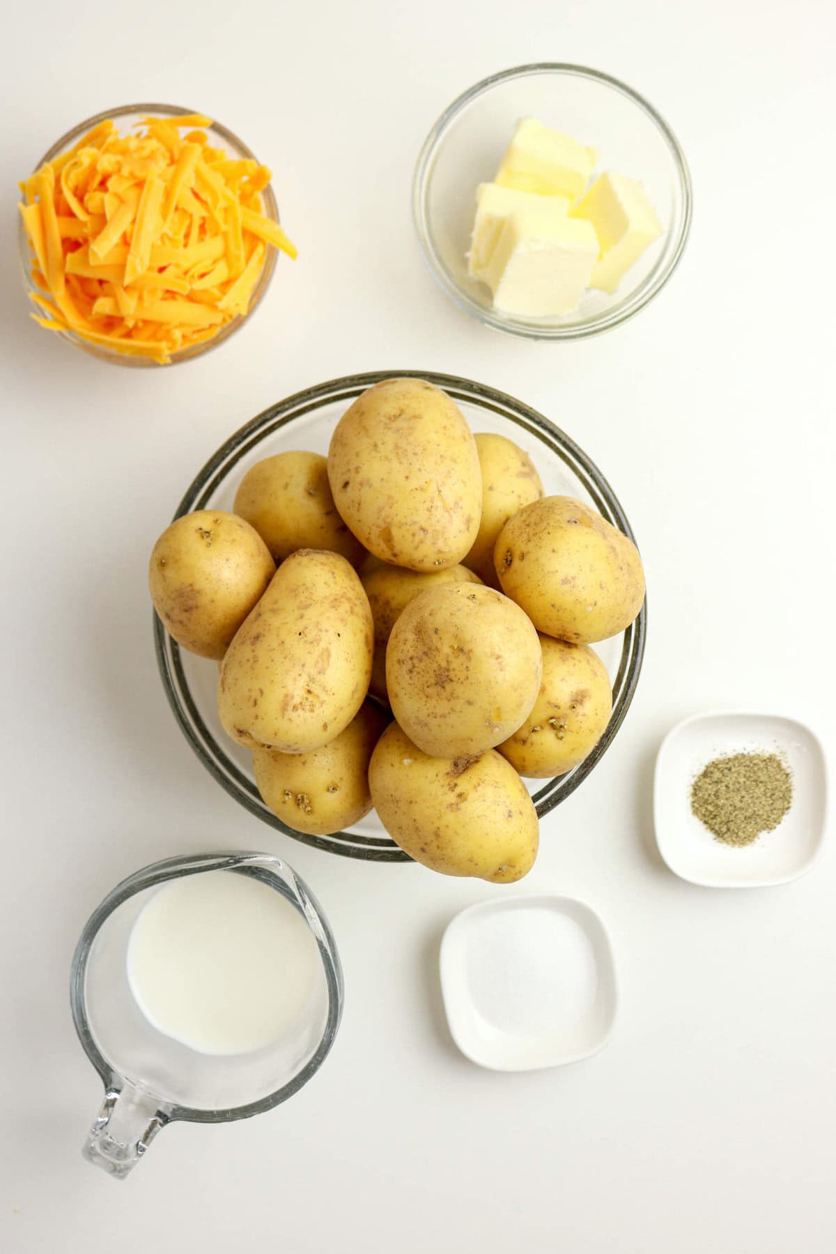 A bowl of whole potatoes is surrounded by bowls of shredded cheddar cheese, butter, milk, salt, and pepper—perfect for making creamy mashed potatoes with cheese on a white surface.