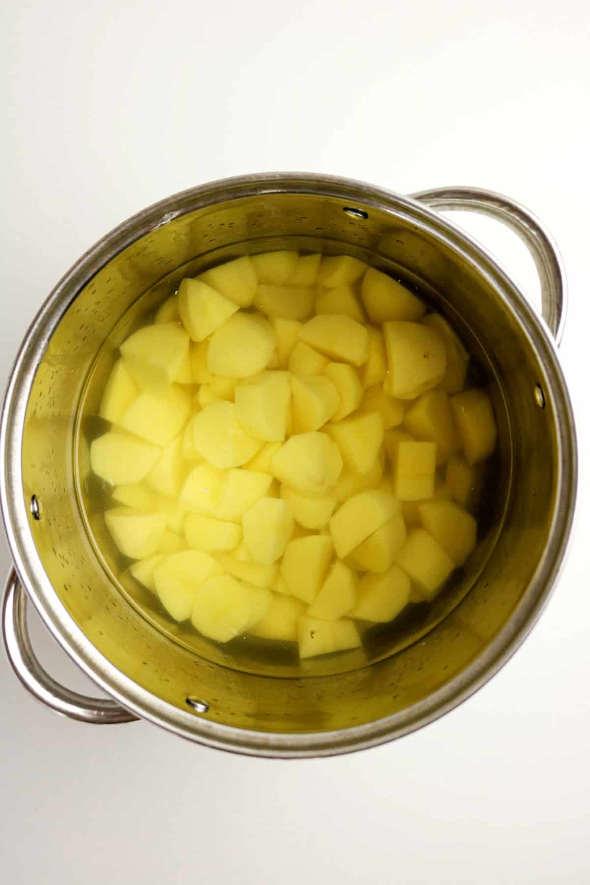A metal pot filled with peeled and chopped potatoes submerged in water, ready to be cooked for creamy mashed potatoes with cheese, viewed from above on a white background.