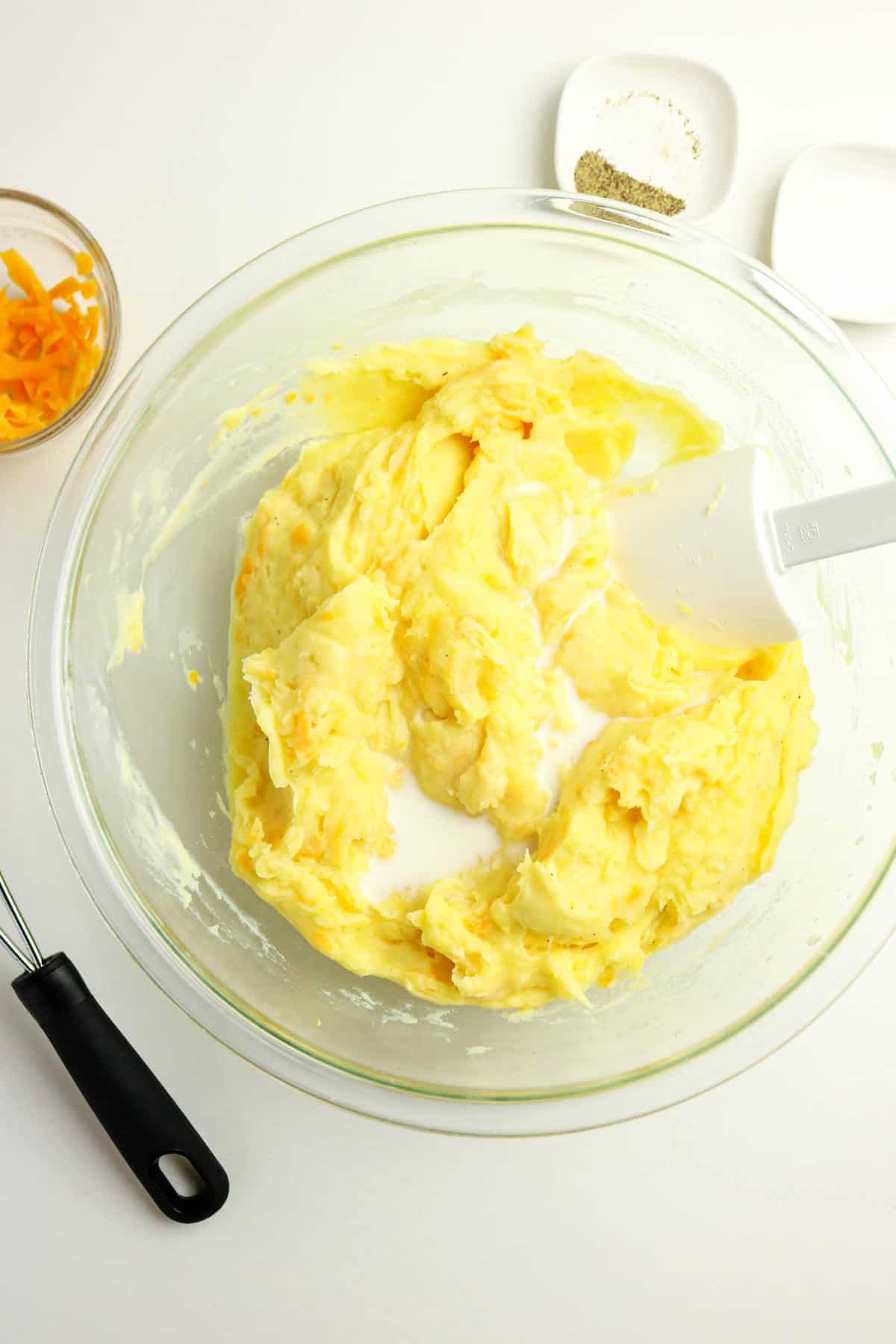 A glass bowl with mashed potatoes being mixed, a white spatula inside, and a splash of milk. Nearby are a small bowl of shredded cheese-perfect for making creamy mashed potatoes with cheese-a whisk, and small dishes with salt and pepper.