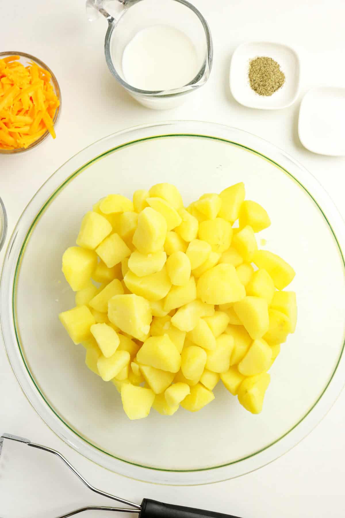 A glass bowl filled with peeled and chopped boiled potatoes, surrounded by small bowls of shredded cheese, milk, black pepper, and salt on a white surface-perfect ingredients for creamy mashed potatoes with cheese. A potato masher is also visible at the bottom.