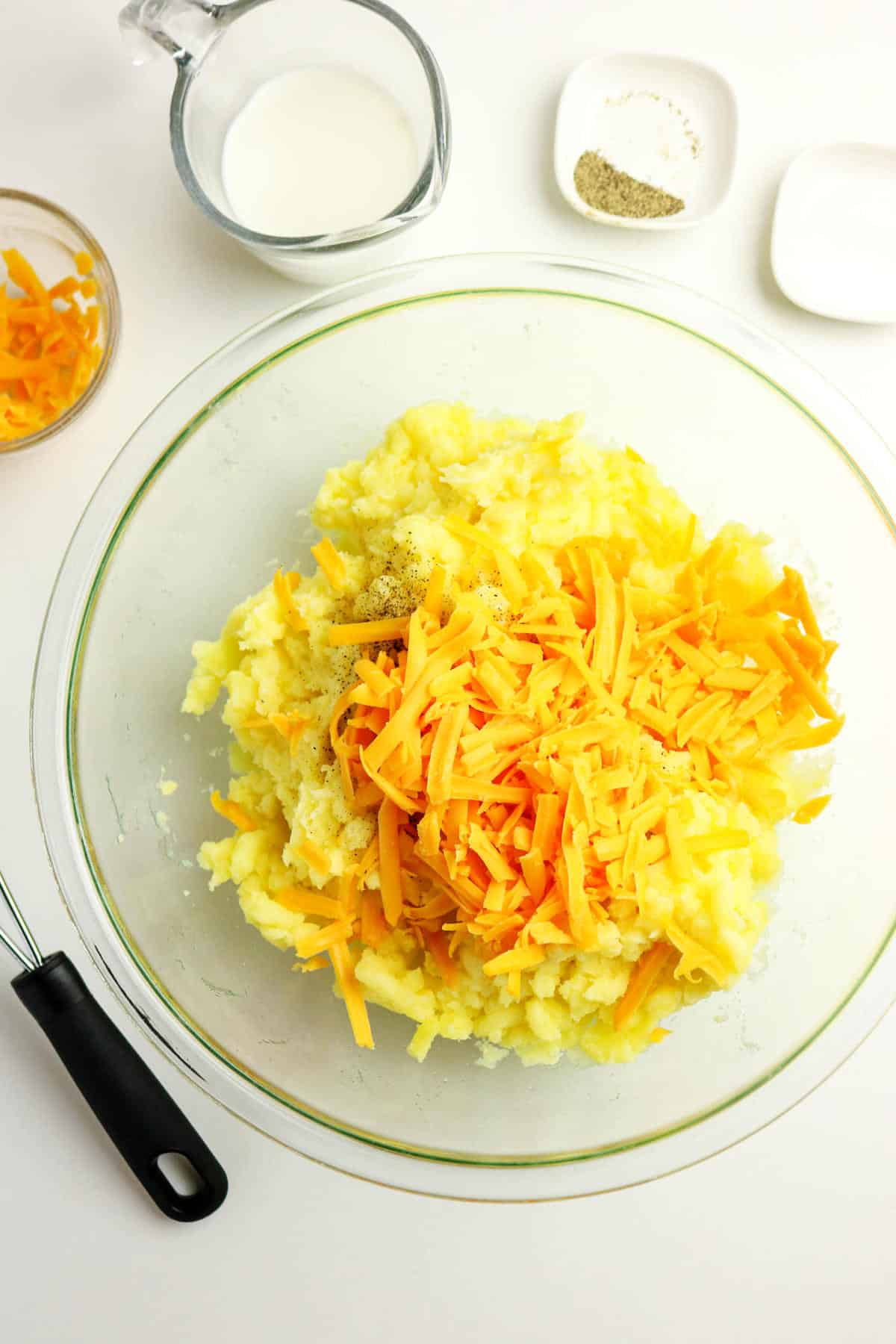 A glass bowl filled with mashed potatoes with cheese, topped with shredded cheddar, sits next to a potato masher. Nearby are a cup of milk, extra shredded cheese, and small plates of salt and pepper for seasoning.