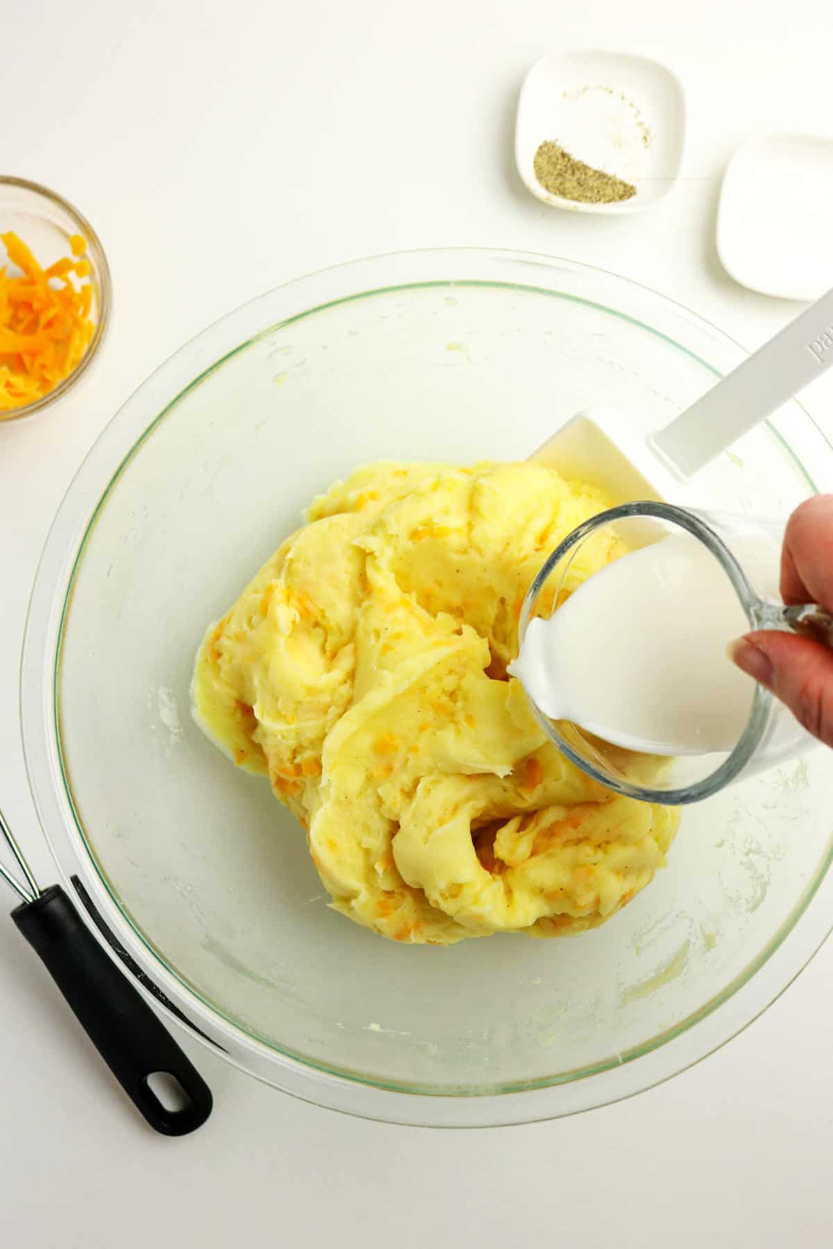 A hand pours milk from a measuring cup into a bowl of mashed potatoes with cheese, with a spatula resting on the side and small bowls of pepper and extra cheese nearby.