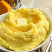 A bowl of creamy mashed potatoes with cheese, topped with melted cheddar and a pat of butter, sits on a light-colored table next to a block of cheddar and a grater.