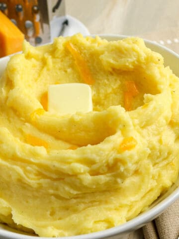 A bowl of creamy mashed potatoes with cheese, topped with melted cheddar and a pat of butter, sits on a light-colored table next to a block of cheddar and a grater.