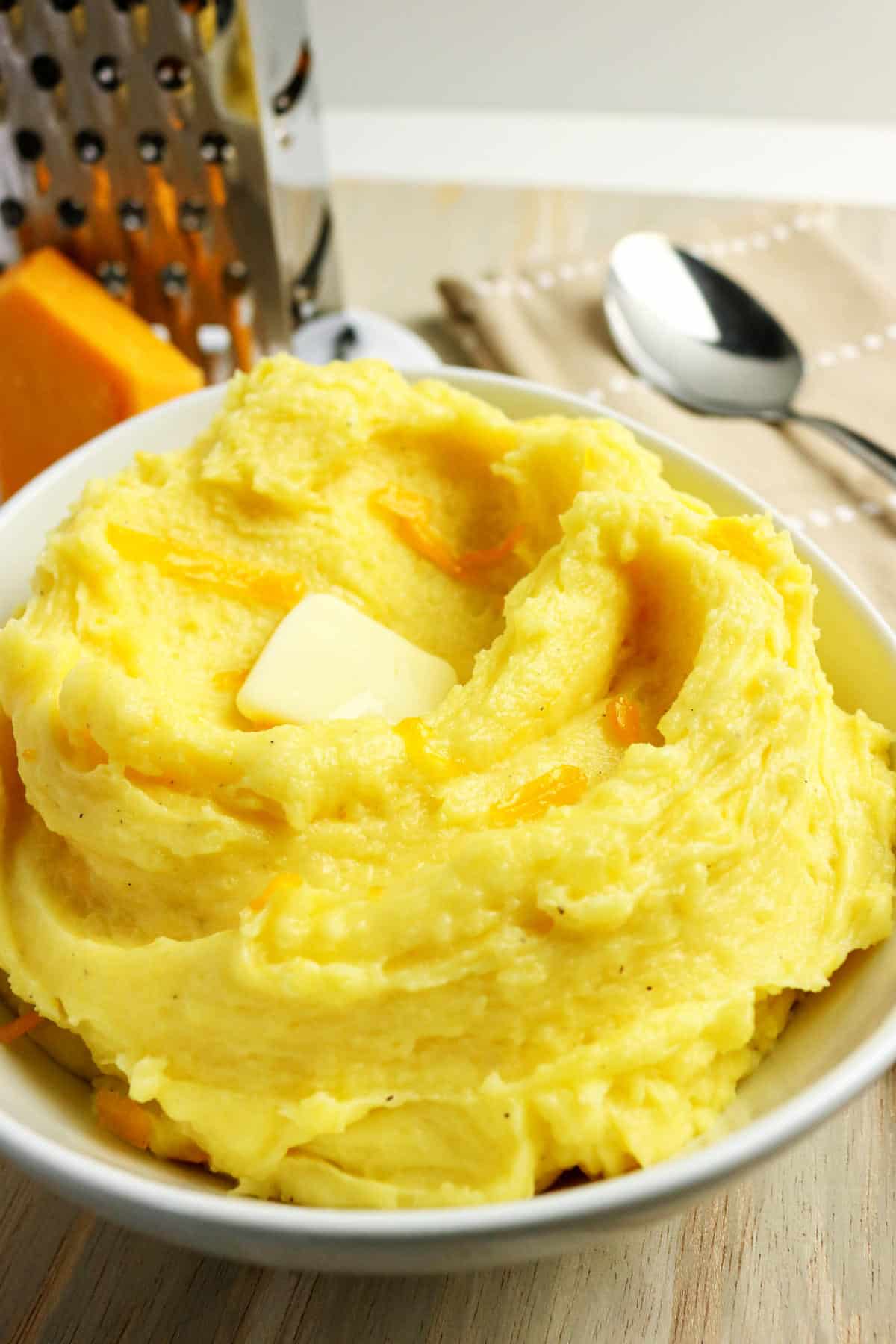 A bowl of creamy yellow mashed potatoes with cheese and a pat of melting butter on top, sitting on a wooden surface near a spoon, napkin, cheese wedge, and a metal grater in the background.