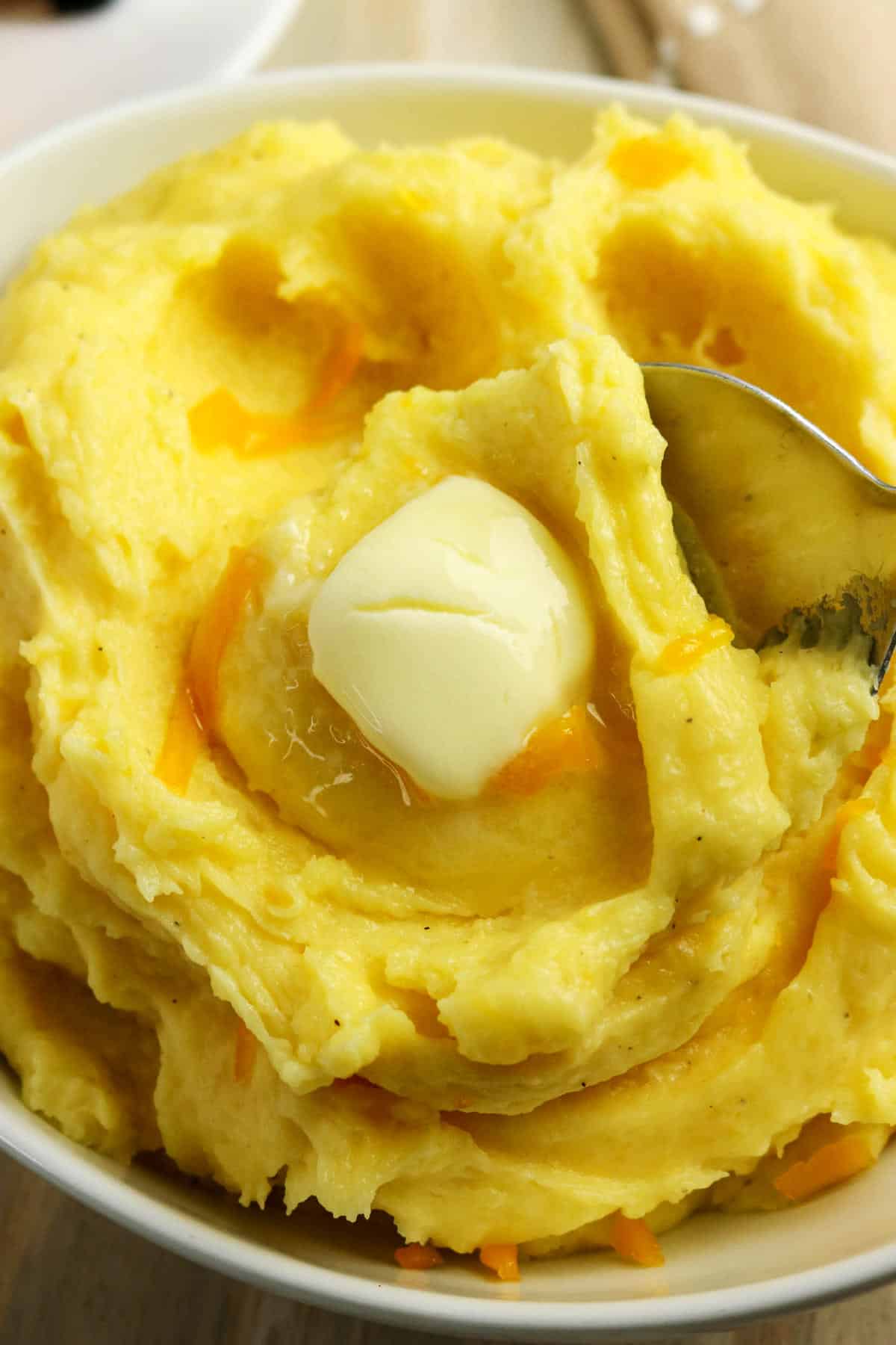 A bowl of creamy mashed potatoes with cheese, featuring a swirl pattern, topped with a melting pat of butter and a spoon scooping into the cheesy potatoes.