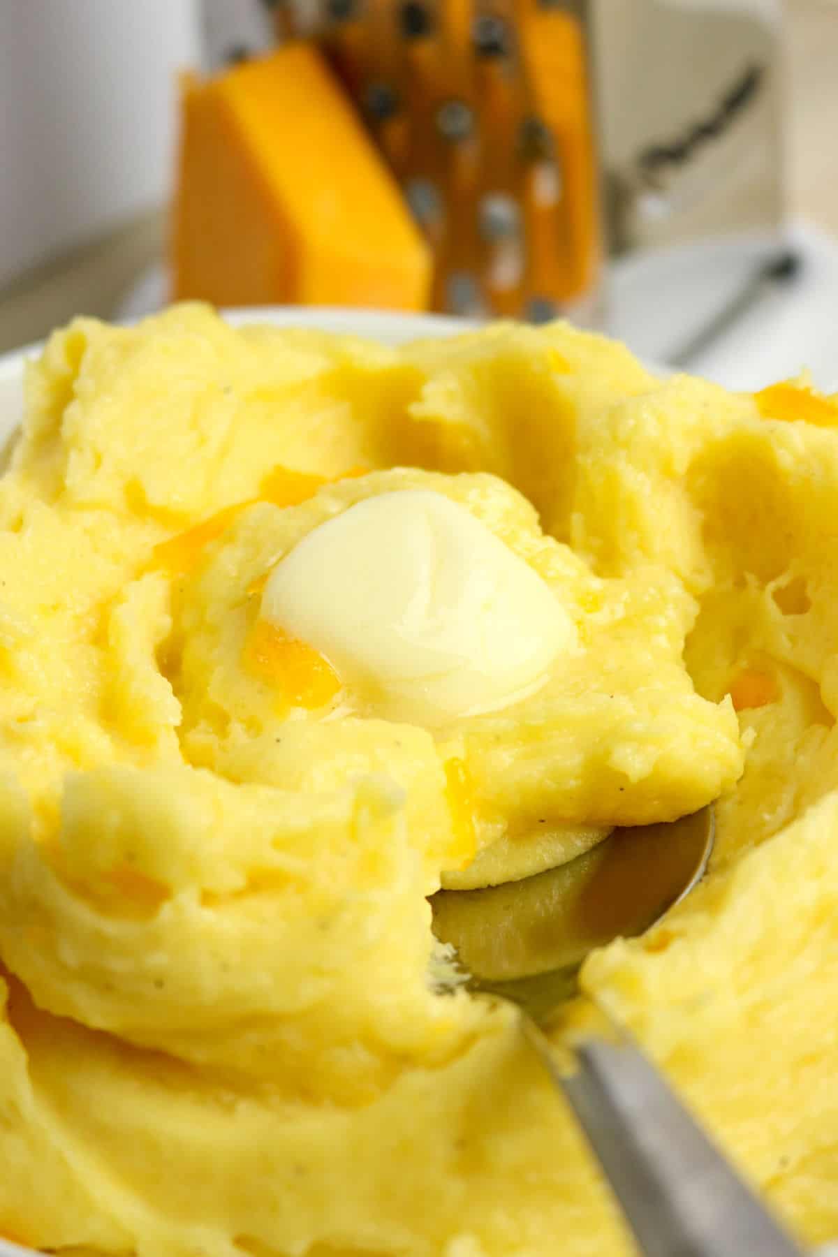 A close-up of creamy mashed potatoes with cheese and melted butter on top, served in a white bowl. A spoon is scooping some out. In the background, there is a block of cheddar cheese and a cheese grater.