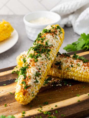 Two ears of Mexican street corn, topped with crumbled cheese, chili powder, and chopped cilantro, rest on a wooden cutting board. Fresh parsley, a bowl of white sauce, and extra corn complete this vibrant Mexican street corn scene.