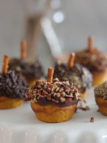 Mini cupcakes and mini pumpkin muffins decorated to look like acorns, topped with chocolate, chopped nuts, and sprinkles, each with a pretzel stick stem, arranged on a white cake stand with a blurred background.