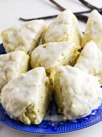 A blue plate holds eight mini vanilla bean scones, each covered in a shiny white glaze and arranged in a circular pattern. The background is white with a few vanilla bean pods visible.