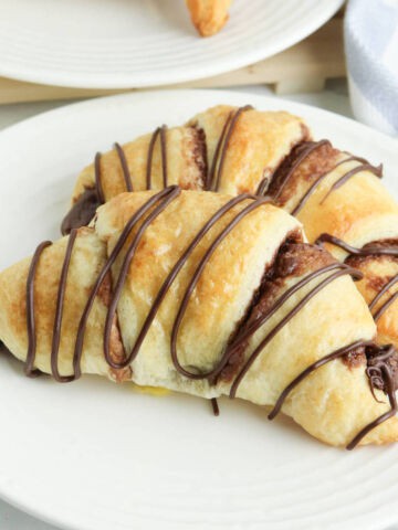 Two Crescent Roll Nutella Croissants, filled and drizzled with rich chocolate, are arranged on a white plate. Another golden croissant is visible in the background on a separate plate.