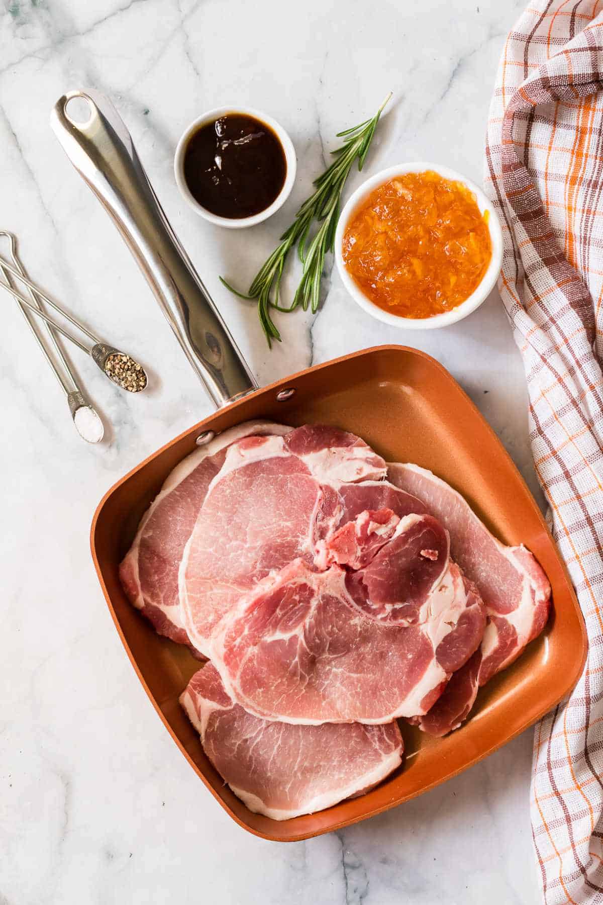 Raw orange pork chops in a copper skillet on a marble surface, surrounded by a bowl of apricot preserves, a bowl of sauce, rosemary, measuring spoons with salt and pepper, and an orange checkered kitchen towel.