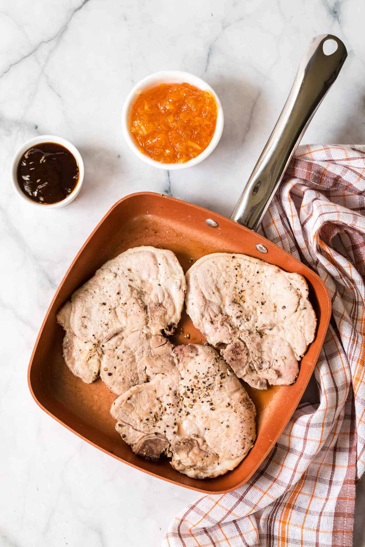 Three cooked orange pork chops in a copper skillet on a marble surface, next to small bowls of orange marmalade and barbecue sauce, with a checkered kitchen towel beside the pan.