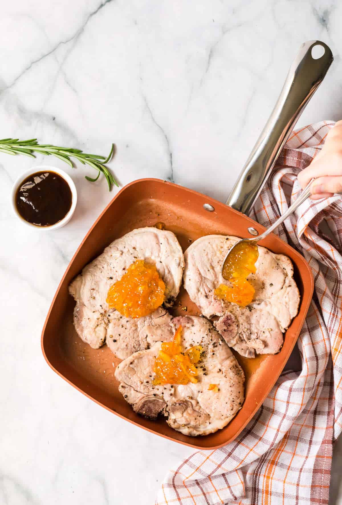 A copper skillet with three orange pork chops, each topped with a dollop of orange marmalade, sits on a marble surface. A hand holds the pan handle with a towel. A bowl of sauce and a rosemary sprig are nearby.