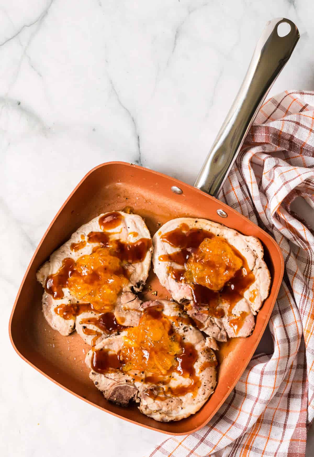 Three cooked orange pork chops in a copper-colored frying pan, topped with a golden-orange sauce and a drizzle of brown glaze. A checkered dish towel rests beside the pan on a marble countertop.