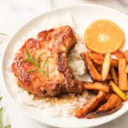 A plate with orange pork chops glazed to perfection, served on white rice, garnished with rosemary, and paired with roasted carrot sticks and a fresh slice of orange.