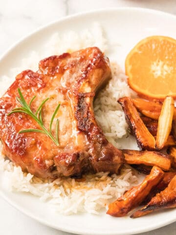 A plate with orange pork chops glazed to perfection, served on white rice, garnished with rosemary, and paired with roasted carrot sticks and a fresh slice of orange.