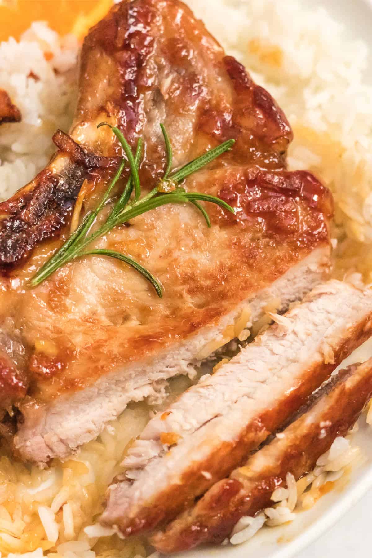Close-up of juicy, cooked orange pork chops garnished with a sprig of fresh rosemary, served over a bed of white rice. Two slices have been cut from the pork, revealing its tender interior.
