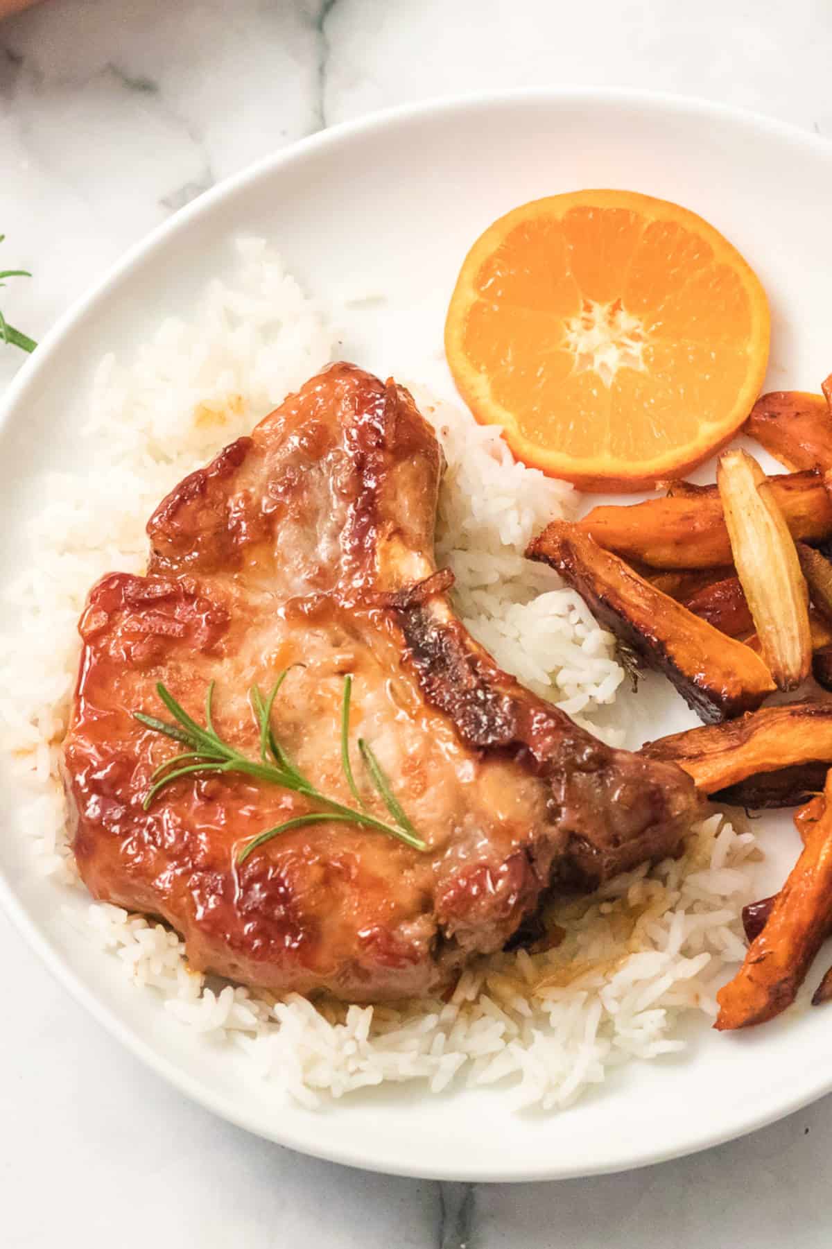 A glazed orange pork chop garnished with rosemary sits on white rice, accompanied by roasted sweet potato fries and a slice of orange on a round white plate.