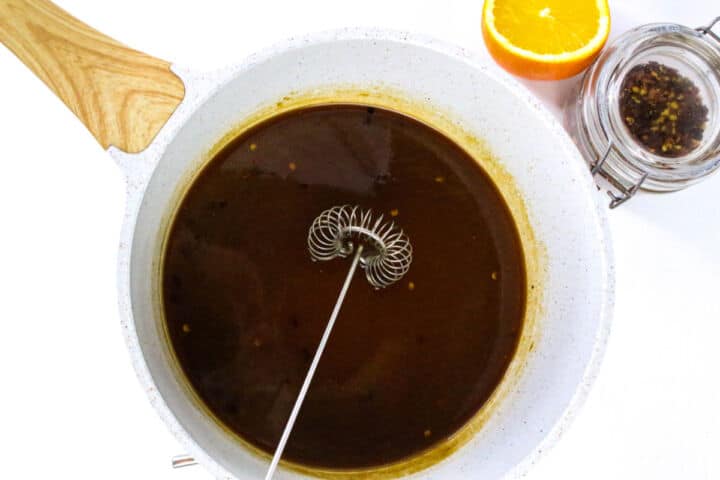 Orange teriyaki sauce simmering in a saucepan with a whisk.