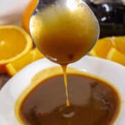 A spoon drizzling golden brown orange teriyaki sauce into a white bowl, with fresh orange slices in the background.