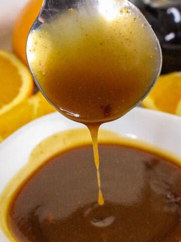 A spoon drizzling golden brown orange teriyaki sauce into a white bowl, with fresh orange slices in the background.