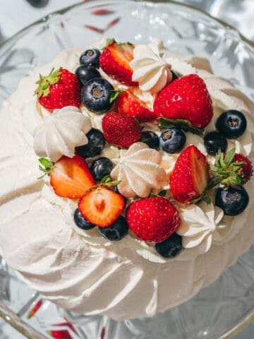 A Berry Pavlova dessert topped with whipped cream, fresh strawberries, blueberries, and small meringues, displayed on a glass cake stand.