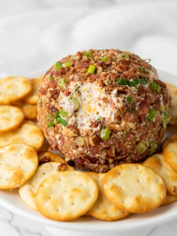 A bacon cheeseball recipe coated with chopped nuts, green onions, and bacon sits on a white plate, surrounded by round crackers. The background is softly blurred with a light-colored cloth.