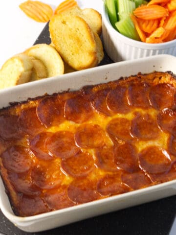 A baked pizza dip casserole topped with melted cheese and pepperoni slices is served in a white dish. Beside it are slices of toasted bread and a bowl of celery and carrot sticks on a black tray.