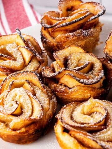 Six Puff Pastry apple roses arranged on a white plate, dusted with powdered sugar. Each pastry features thin apple slices spiraled to resemble blooming roses. A red and white striped cloth sits in the background.