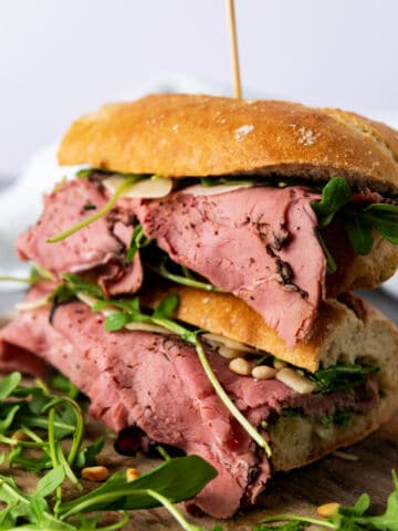 A sandwich filled with thick slices of roast beef, fresh arugula, and pine nuts, stacked on a wooden board. The sandwich is cut in half and held together with a wooden skewer.