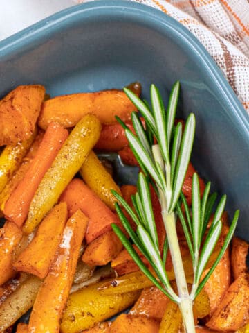 A close-up of roasted parsnips and carrots in a blue dish, garnished with a fresh sprig of rosemary. A white and orange checkered cloth is partially visible in the background.