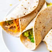 Two flour tortillas filled with cheddar cheese, guacamole, cilantro, and Instant Pot shredded chicken are arranged side by side on a white plate for delicious Instant Pot Shredded Chicken Tacos.