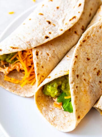 Two flour tortillas filled with cheddar cheese, guacamole, cilantro, and Instant Pot shredded chicken are arranged side by side on a white plate for delicious Instant Pot Shredded Chicken Tacos.