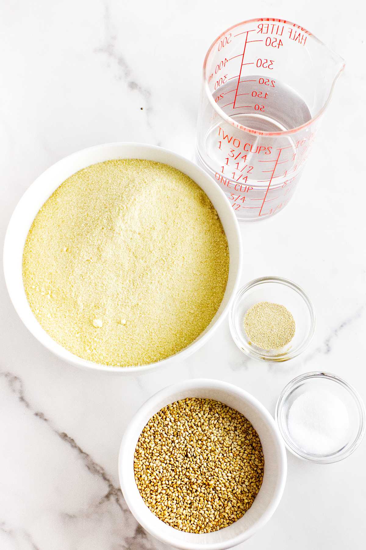 A measuring cup of water, a bowl of cornmeal, a small bowl of millet, a dish of salt, and a small dish of yeast are arranged on a white marble surface—ingredients often used in Moroccan semolina bread.