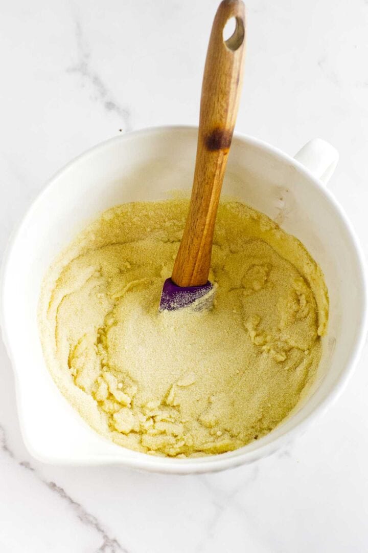 A white mixing bowl with yellow cornmeal, perfect for making Moroccan semolina bread, and a wooden spoon inside, sits on a marble surface.