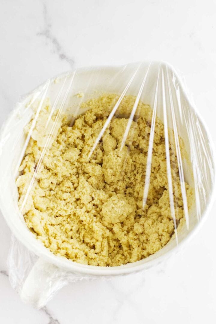 A bowl covered with plastic wrap containing yellow crumbly dough for Moroccan semolina bread sits on a white marble surface.