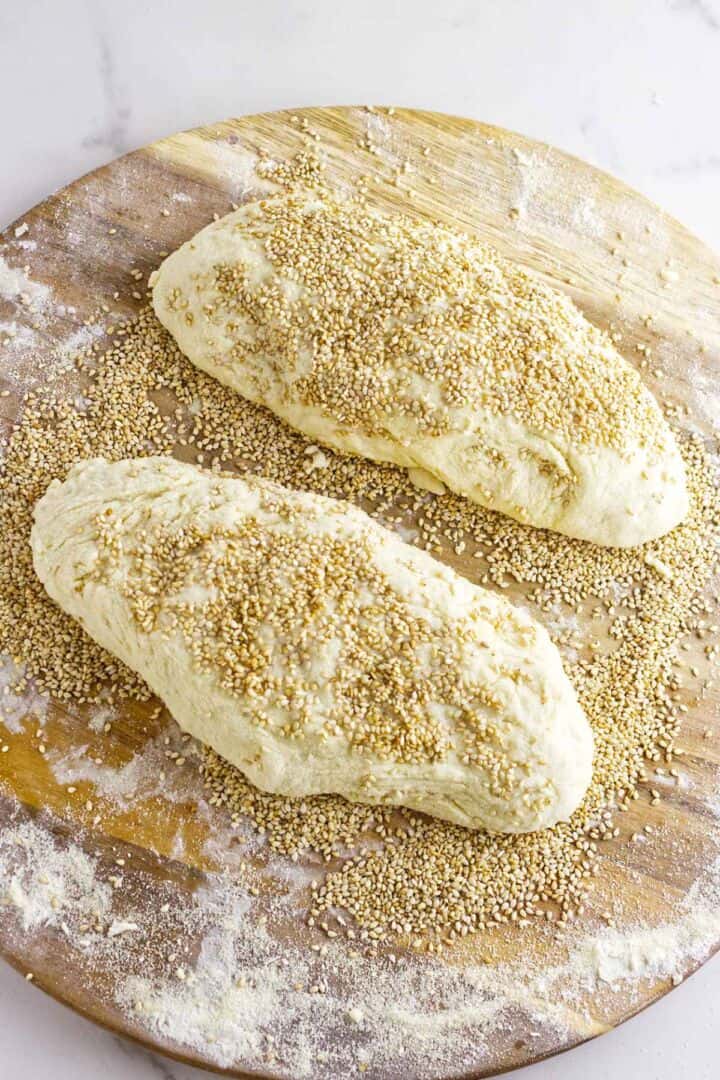 Two oval-shaped pieces of Moroccan semolina bread dough topped with sesame seeds rest on a floured wooden surface, with extra sesame seeds scattered around them.