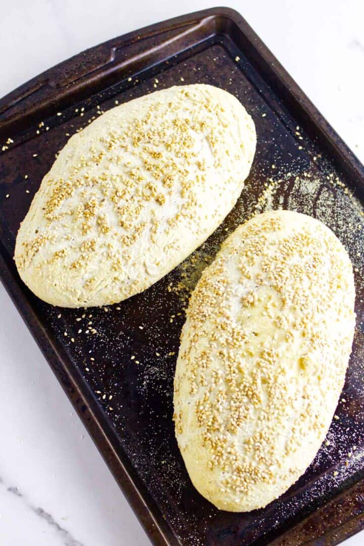 Two oblong loaves of Moroccan semolina bread sprinkled with sesame seeds rest on a dark baking sheet, with scattered flour and seeds around them. The background is a white surface.