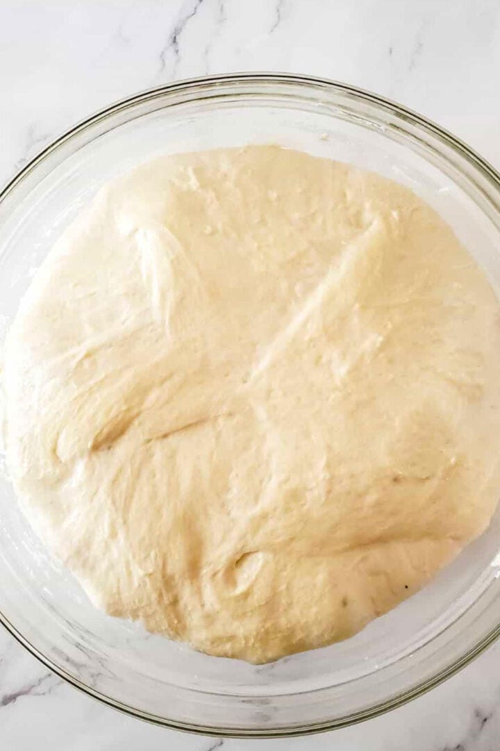 A glass bowl filled with risen, fluffy Moroccan semolina bread dough sits on a white marble surface.