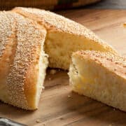 A round Moroccan semolina bread with a golden crust and sesame seeds on top sits on a wooden surface. A wedge-shaped piece has been cut out, revealing the soft, fluffy interior.