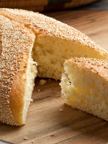 A round Moroccan semolina bread with a golden crust and sesame seeds on top sits on a wooden surface. A wedge-shaped piece has been cut out, revealing the soft, fluffy interior.