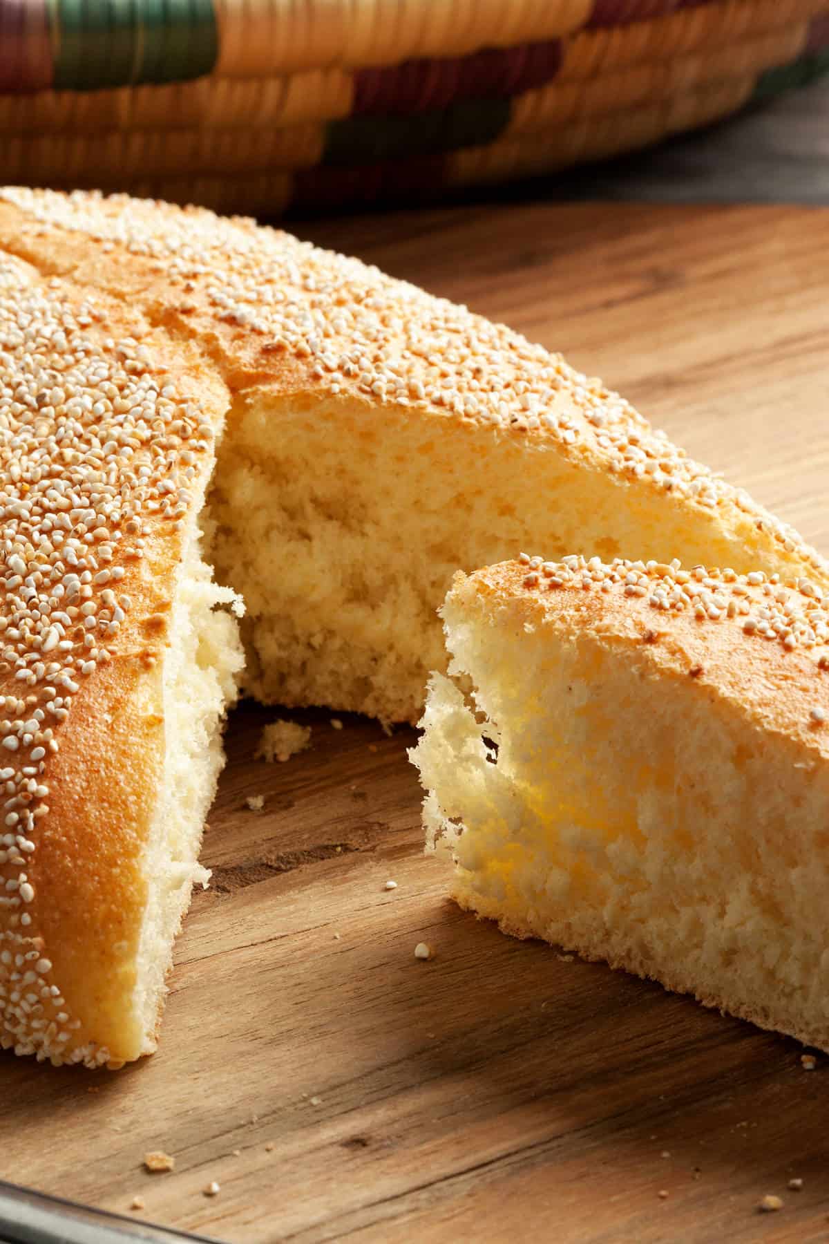 A close-up of a round, fluffy Moroccan semolina bread loaf with a slice cut out, showing its soft interior and golden crust topped with sesame seeds, resting on a wooden surface.