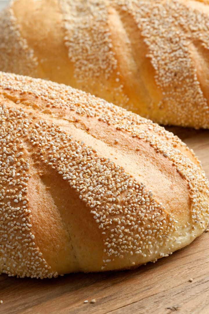 Two loaves of freshly baked Moroccan semolina bread with golden crusts, topped with sesame or poppy seeds, rest on a wooden surface. The bread features signature diagonal slashes on top.