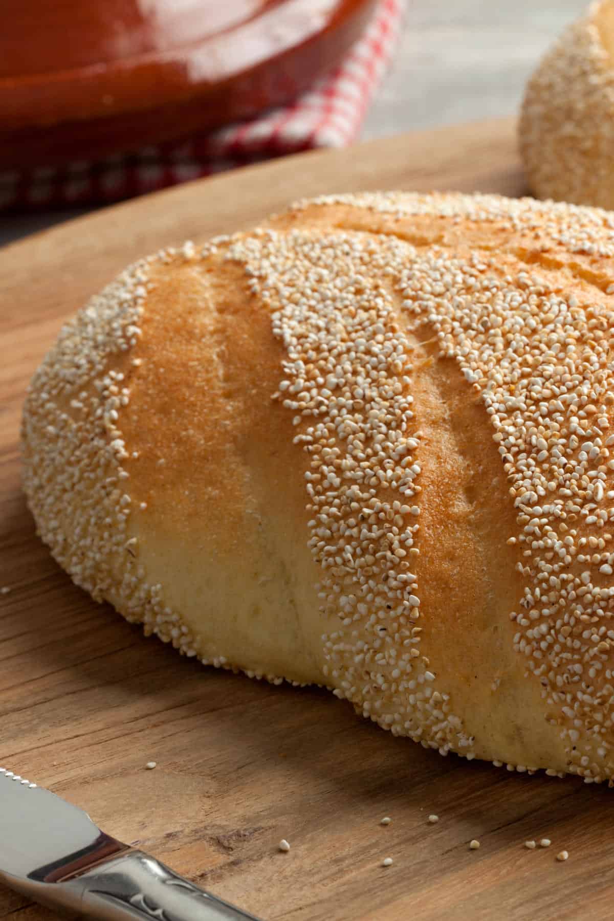 A close-up of Moroccan semolina bread covered with sesame seeds, resting on a wooden cutting board with a butter knife nearby. A red-checkered cloth is visible in the background.
