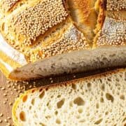 A loaf of Moroccan semolina bread topped with sesame seeds sits on a wooden surface, with one slice cut to reveal its airy, golden crust and soft, open crumb. Sesame seeds are scattered around the bread.