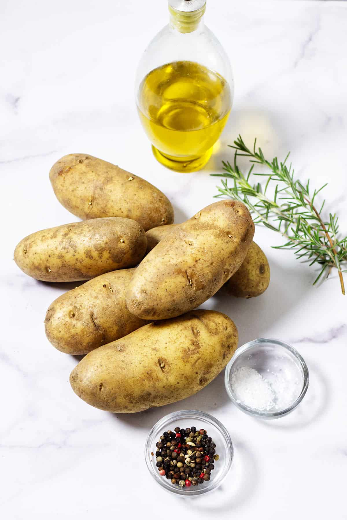 Six russet potatoes on a white surface, next to a glass bottle of olive oil, a sprig of rosemary, and small bowls of mixed peppercorns and salt—perfect ingredients for delicious roasted rosemary potatoes.
