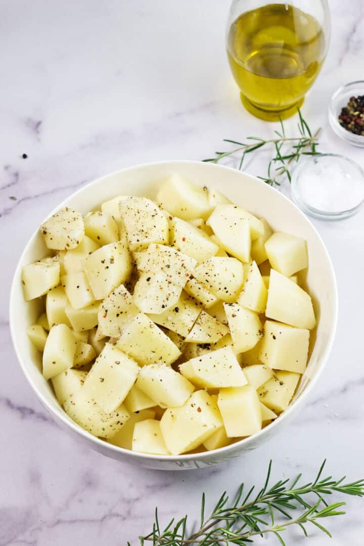 A white bowl filled with peeled, cubed potatoes seasoned for roasted rosemary potatoes sits on a marble surface. Nearby are a sprig of rosemary, a small bowl of salt, a glass container of oil, and a pepper grinder.